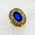 Vintage gold tone Belle Époque style ring with oval sapphire blue glass center stone, paste halo, and black enamel carved detailing