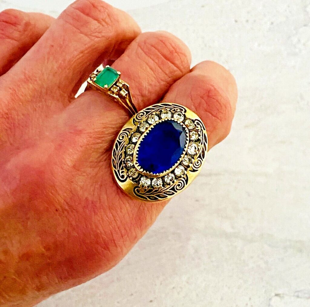 Vintage gold tone Belle Époque style ring with oval sapphire blue glass center stone, paste halo, and black enamel carved detailing