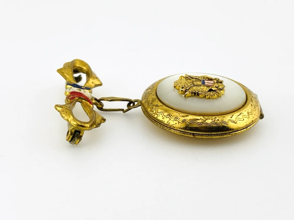 Gold-plated WWII sweetheart locket brooch with an eagle-and-shield emblem on mother of pearl, engraved border, and red, white, and blue enameled bow.