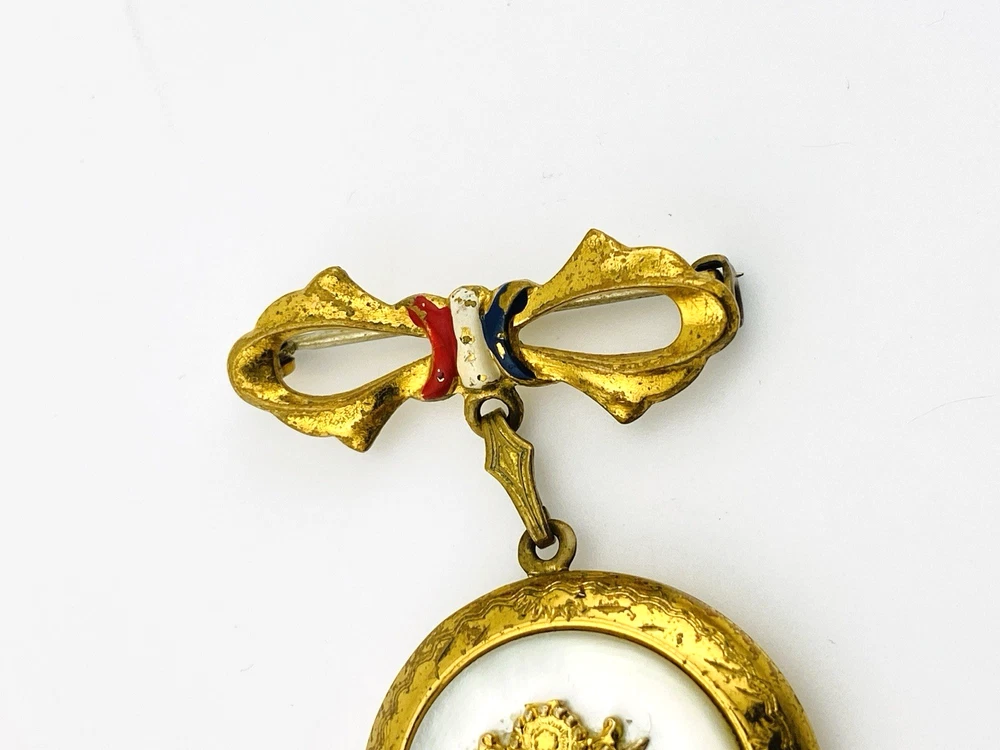 Gold-plated WWII sweetheart locket brooch with an eagle-and-shield emblem on mother of pearl, engraved border, and red, white, and blue enameled bow.