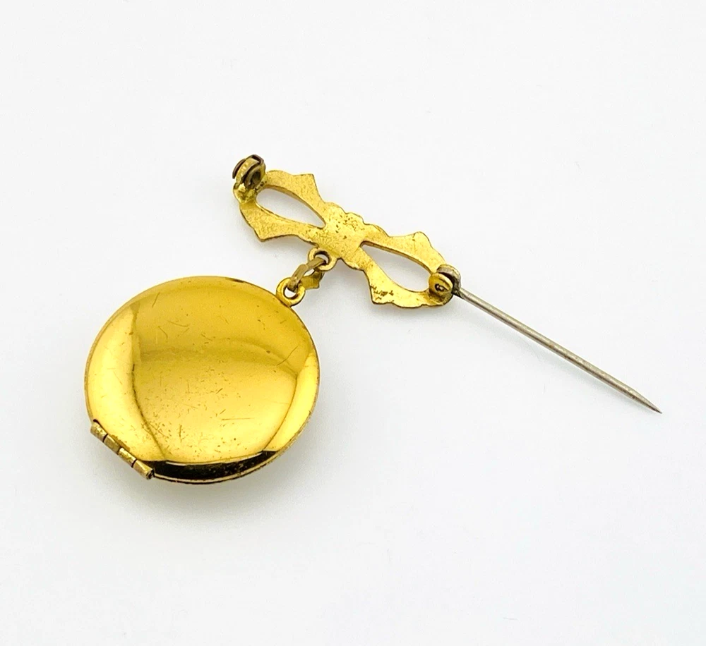 Gold-plated WWII sweetheart locket brooch with an eagle-and-shield emblem on mother of pearl, engraved border, and red, white, and blue enameled bow.