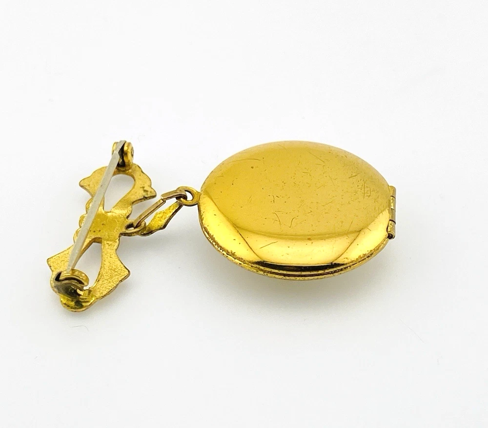 Gold-plated WWII sweetheart locket brooch with an eagle-and-shield emblem on mother of pearl, engraved border, and red, white, and blue enameled bow.